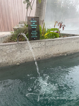 A water fountain launches fresh, cold spring water into the hot lagoon. A sign in Icelandic and english says "Please remember to stay hydrated"