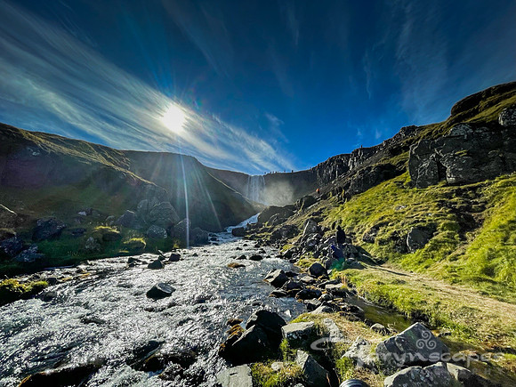 A cascade falls between two moss covered vocanic rock cliffs. Deep blue sky is above and a bright starburst sun washes out some of the color