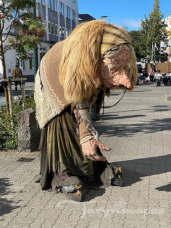 Large hunched over troll woman with a pointy nose sculpture stands on the sidewalk.