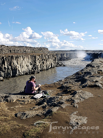 I sit crosslegged at the cliff edge of a river, with my hat on backwards looking at my camerabag. In the distance the waterfall I want to shoot.