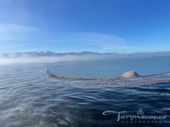 The stone edge of an infinity pull shimmers below the water's surface. Past the edge lies the ocean, blue skies and mountains