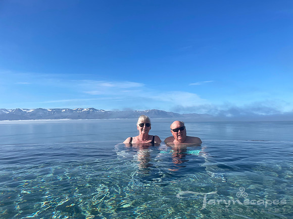 An older couple sit at the edge of an infinity pool and the ocean behind them. In the distance, snowcapped mountains.