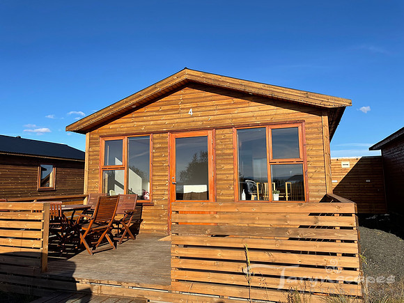 A wood cabin with 2 large windows on either side of the glass front door and a front porch with a table and chairs