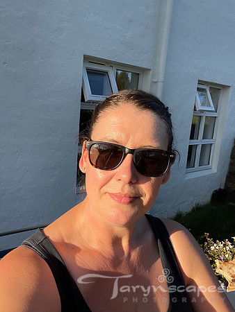 A selfie with my dark hair pulled back, sunglasses on, black tank top with the sun brightly shining on my face.
