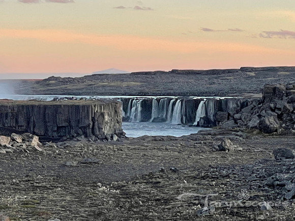 A rocky foreground leads up to a very wide waterfall in the distance flowing down a cliff withink and yellow skies