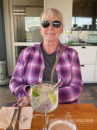 Mom with white hair pulled back with a bun, bangs and aviator glasses. A gin & tonic sits in front of her as she smiles.
