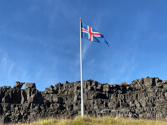Black cliffs loom behind an Icelandic flag blowing in wind in front of bright blue sky.