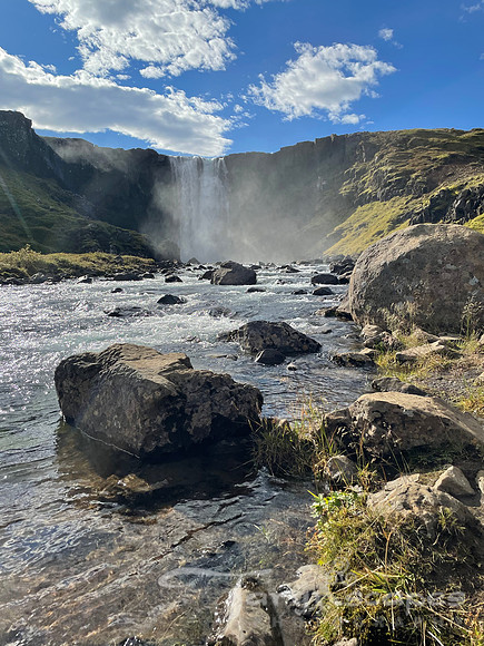 Waterfall illuminated by the bright sun, mist obscuring the details. Above bright blue sky with puffy clouds
