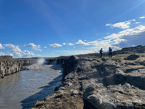 Harsh landscpae of rock drops off into a cliff and waterfall in the distance. Two small outlines of people show the scale of the waterfall