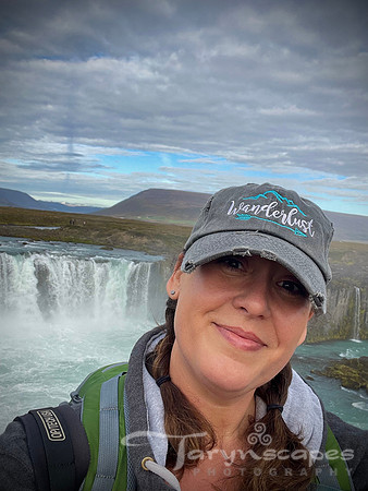 My smiling face with my hair in braids and my Wanderlust hat in front of Godfoss waterfall