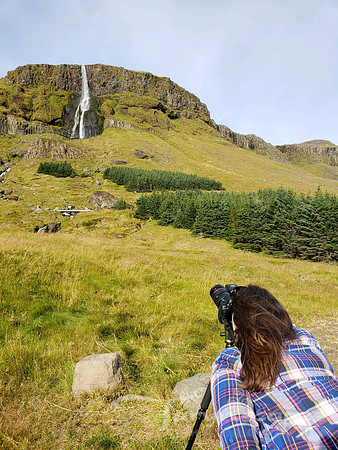 I'm leaning over my camera on it's tripod while I shoot a tall skinny waterfall coming out of a mountain in the distance.