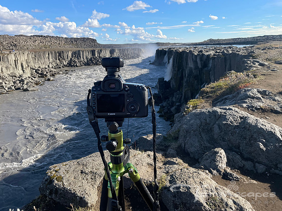 A DSLR Canon camera sits on a green tripod framed between the two cliffs pointing at a waterfall