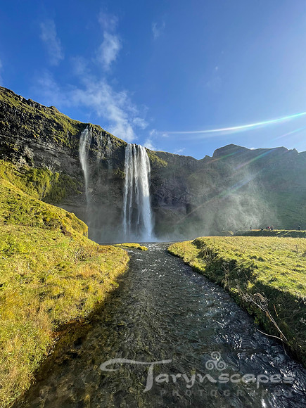 A stream leads up to a tall watefall lit by the sunshine.