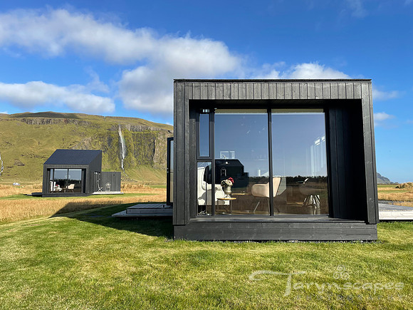 Moddern square cabin with black siding and full length windows in the back.