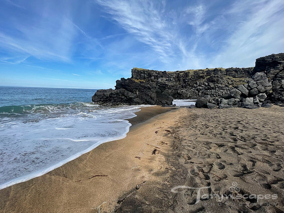 An ocean wave slides up the beach. The sand changes from a golden to dark brown. In the distance a black rock structure stratles the beach and the ocean